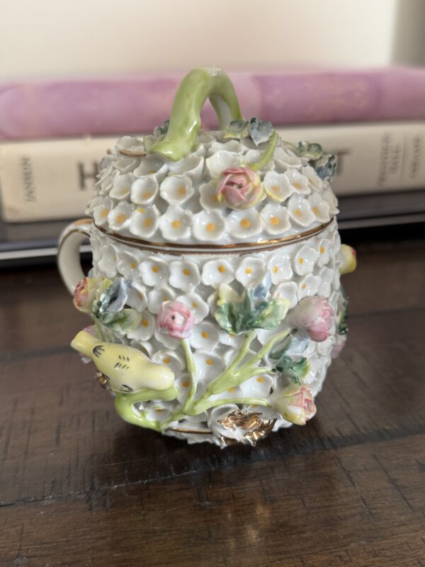 The Meissen Schneeballen Cup is a delicate white porcelain piece with raised flower designs, pink roses, a green stem-shaped handle, a yellow bird, and intricate floral details, displayed on wood with blurred books in the background.