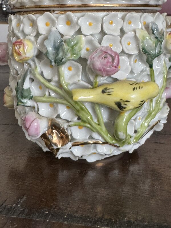Close-up of a Meissen Schneeballen Cup, adorned with raised white flowers with yellow centers, pink rosebuds, green leaves, and a yellow bird among foliage. Gold accents add elegance to this decorative porcelain piece displayed on wood.