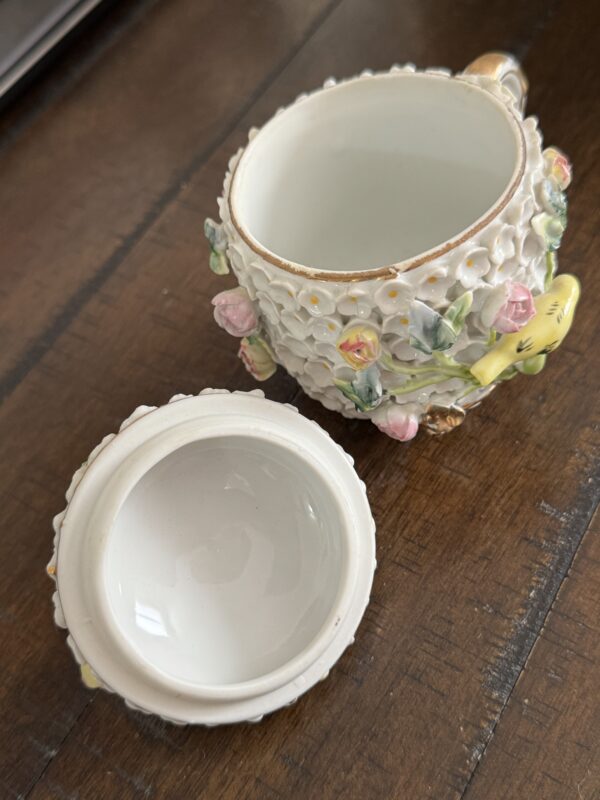 The Meissen Schneeballen Cup is a white porcelain container with a removable lid, featuring textured white flowers, pastel roses, green leaves, and a yellow butterfly. It sits elegantly on a wooden surface.