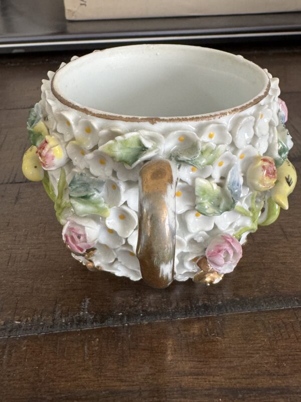 The Meissen Schneeballen Cup, a white ceramic piece with a gold handle and raised floral designs—pink roses, green leaves, yellow flowers—rests empty on a wooden table, showcasing its ornate and textured charm.