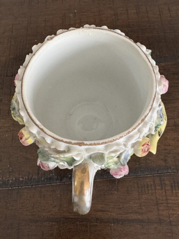 A top-down view of the Meissen Schneeballen Cup, featuring a gold handle, floral patterns, and raised flower motifs on its exterior. The cup rests on a wooden table, with visible wear around the rim inside.