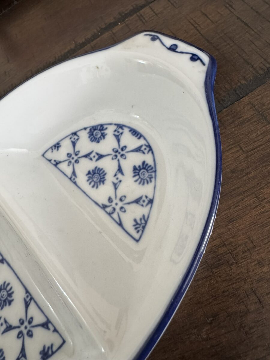A close-up of a white ceramic dish with blue floral patterns and blue trim, resting on a wooden surface. The dish has a divided section and a small handle with blue accents. The wood underneath shows visible grain and texture.