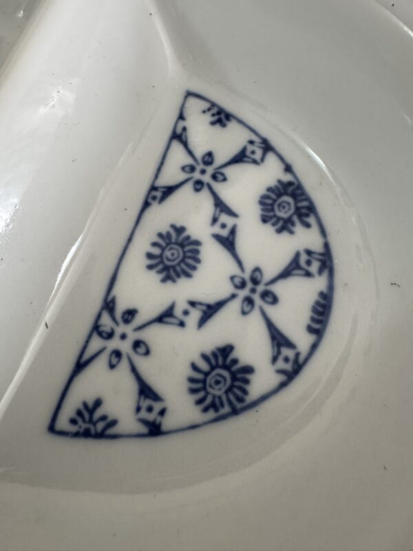 A close-up of a white ceramic surface featuring a half-moon-shaped blue floral and geometric pattern, with various intricate designs and symmetrical motifs. The surrounding area is glossy and smooth, reflecting light.