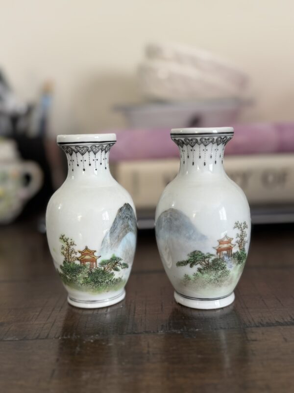 Two small white porcelain vases with painted landscapes, including mountains, trees, and red pagodas. The vases have black decorative rims and sit on a dark wooden surface with a blurred background of books and household items.