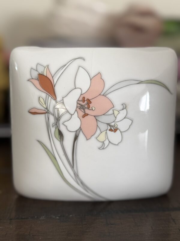 A white ceramic cup with a delicate design of pink and white flowers and green leaves, outlined in thin gray lines, is shown. The background is blurred, keeping the focus on the floral artwork on the cups smooth surface.