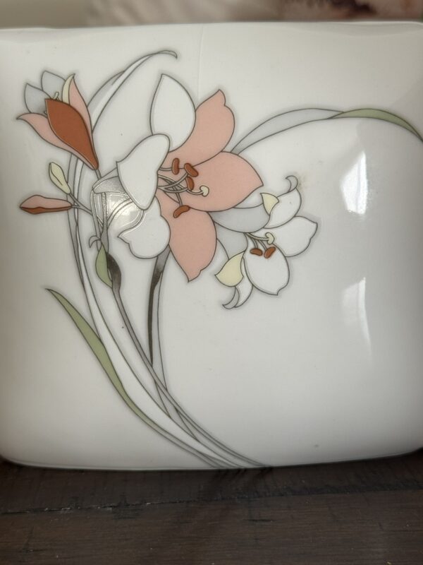 A close-up of a white ceramic surface features a delicate floral design with pink, white, and orange flowers, green leaves, and thin gray stems, all outlined in fine lines. The flowers resemble lilies and the background is glossy and smooth.