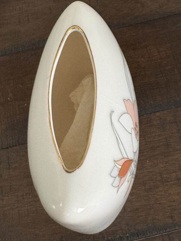 A cream-colored ceramic napkin holder shaped like a rounded teardrop sits on a wooden surface. It has a large oval opening edged in gold and a floral design with pink and orange flowers on one side.