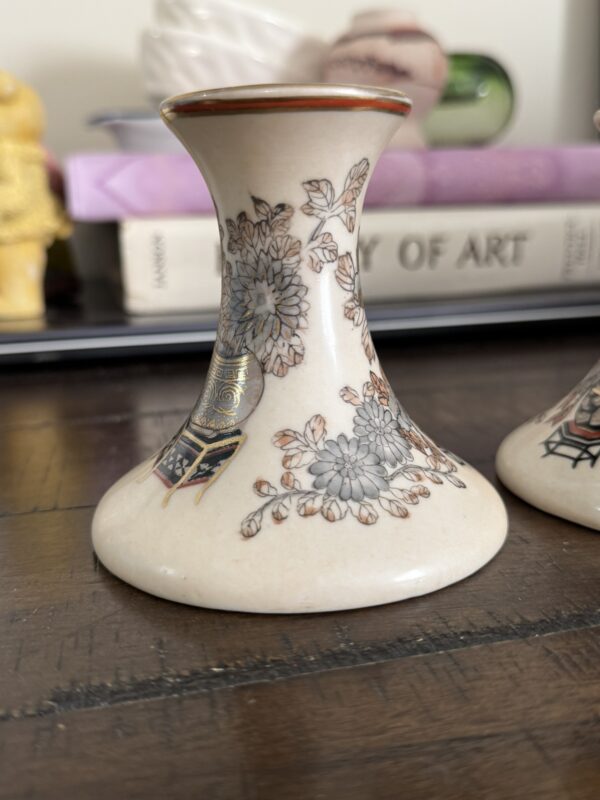 A small, flared ceramic vase with a wide base and narrow neck, decorated with intricate floral patterns in gray and gold. The vase sits on a wooden surface, with blurred books and other objects in the background.