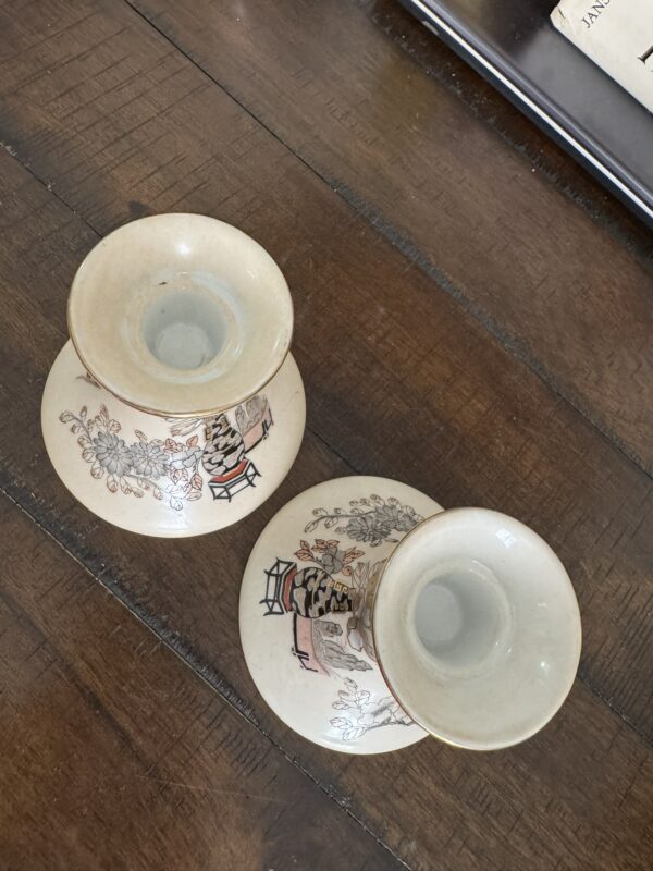 Two vintage cream-colored ceramic candlestick holders with oriental-inspired floral and figure designs sit on a wooden table. The holders feature hand-painted patterns in muted red, green, and black. A book and tray are partially visible at the edge of the image.