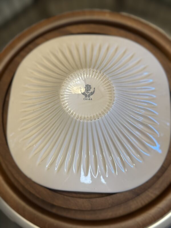 A white ceramic dish is turned upside down, revealing a textured, radiating pattern and a stamp that reads “China” with a small emblem above it. The dish rests on a round wooden surface with a raised edge. The background is softly blurred.