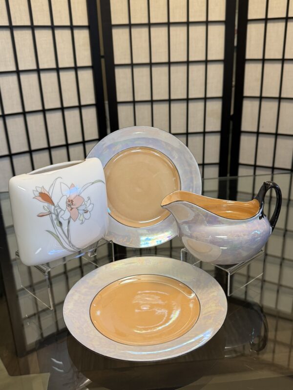 Four pieces of vintage china, including two round plates, a square plate with orange and white floral design, and a matching cream pitcher with a dark handle, are displayed on glass with a black shoji screen backdrop.