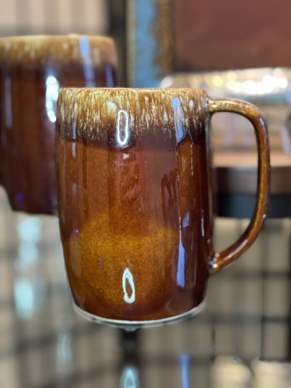 A close-up of a glossy, brown ceramic mug with a large handle, featuring a gradient from dark brown at the top to a lighter amber shade at the bottom. The mug has a shiny glaze with reflective highlights and is set against a blurred background.