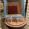 A square ceramic plate with a glossy brown center and textured blue edges featuring swirl patterns stands upright on a clear plastic display easel, placed on a round wooden base. The background includes a glass tabletop and folding screen.