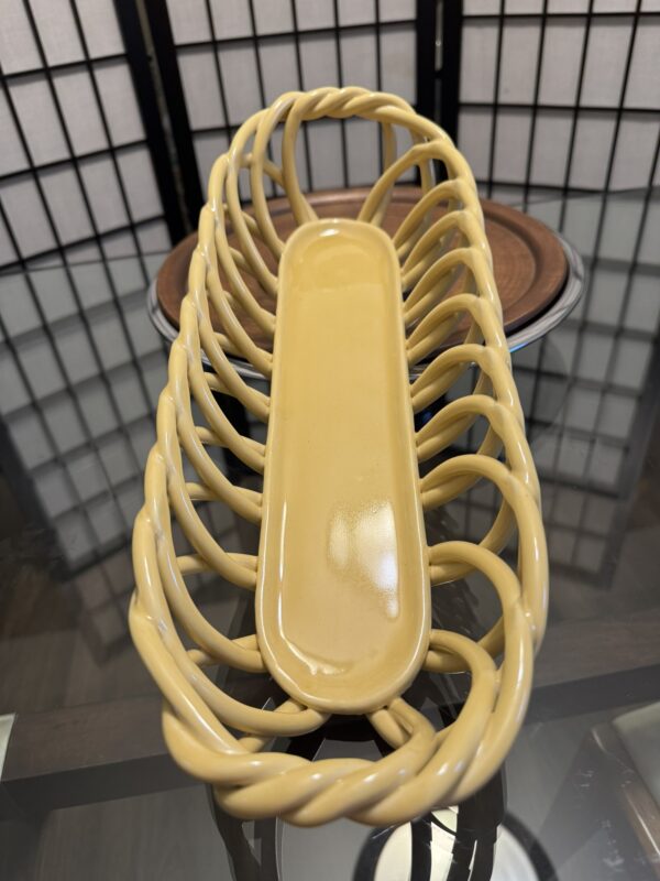 A yellow ceramic bread basket with an open, twisted lattice design sits on a glass table. Behind it are stacked brown plates and a black folding screen with rectangular white panels, reflected in the tabletop.