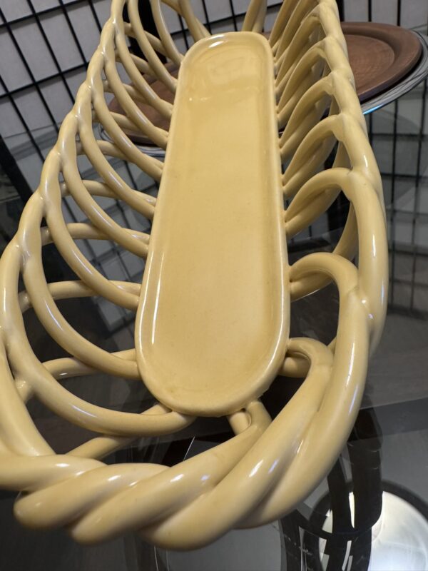 A long, yellow ceramic bread basket with a twisted, open-lattice design sits on a glass table. A round wooden tray and a blue-rimmed plate are partially visible in the background. The setting appears indoors, with grid-like window panels behind.