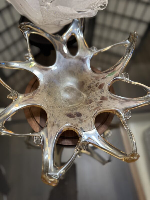 A clear, glass decorative bowl with an organic, starburst shape and multiple uneven, curved protrusions sits on a wooden pedestal. The glass has an iridescent finish and is displayed indoors, with blurred background details.