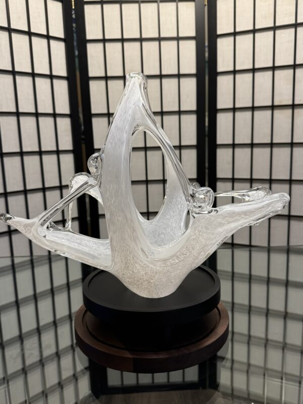 A clear abstract glass sculpture with flowing, curved shapes sits on a round black and wooden base. The background features a folding screen with a grid pattern and white panels, reflected on the shiny surface beneath the sculpture.