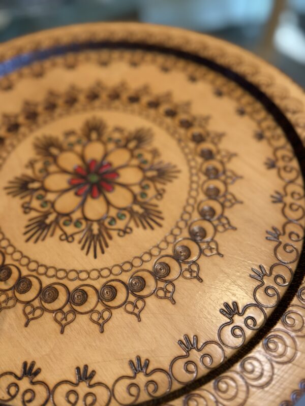 A close-up of a round, light wooden tray features intricate, symmetrical floral and geometric patterns burned into the surface, with small accents of red and green at the center. The designs form concentric circles, giving the tray an ornate, decorative look.