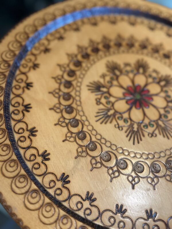 A close-up of a round wooden table shows intricate, symmetrical floral and geometric patterns etched and burned into the surface, with a central flower design highlighted by touches of red and green. The outer rim features repeating spiral and heart motifs.