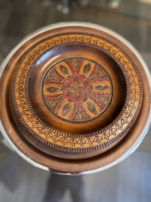A round wooden plate with an intricate, colorful floral and leaf pattern in red, orange, and yellow at the center, surrounded by a textured, carved border. The plate rests on a plain wooden platter, both placed on a glass surface.