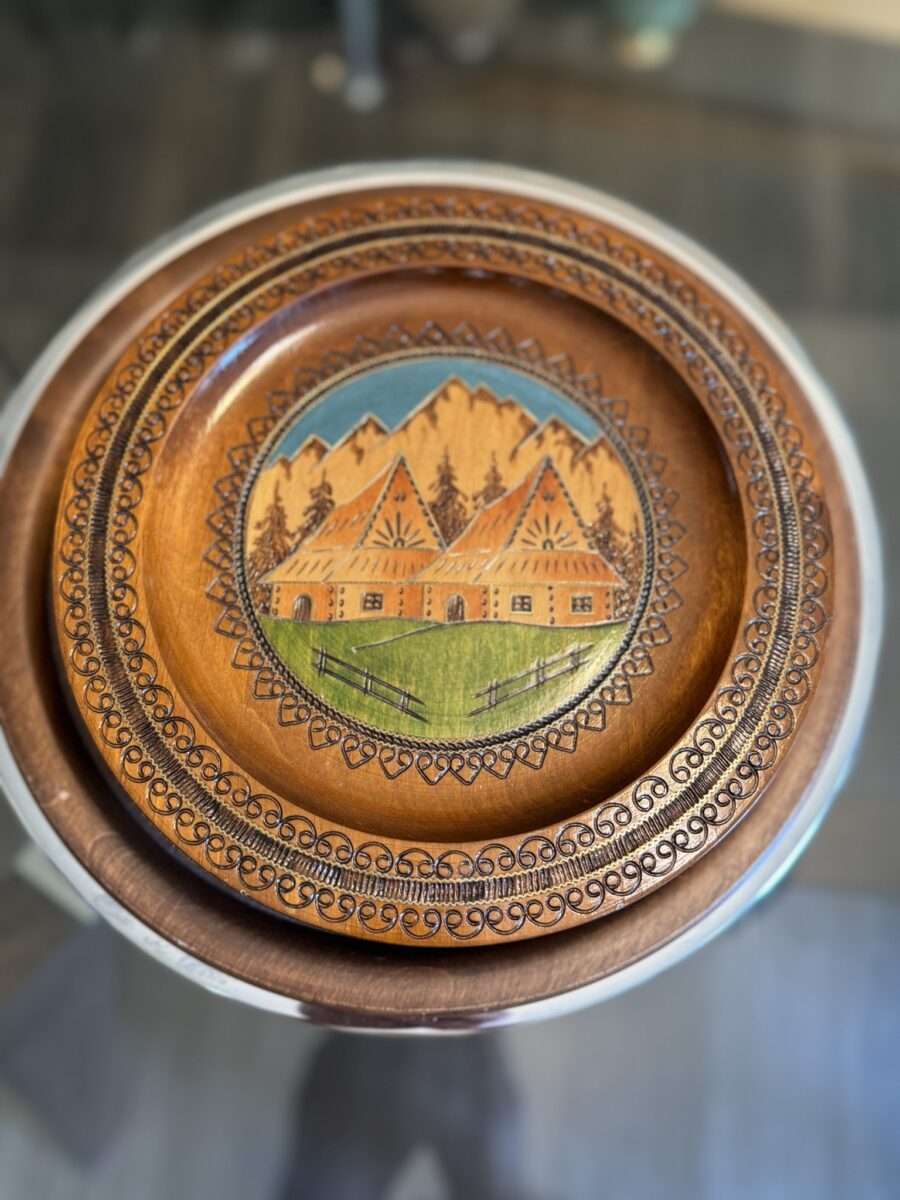 A round, decorative wooden plate with intricate carved borders, featuring a painted scene of three cottages with red roofs, green lawns, and a background of tall brown mountains under a blue sky. The plate sits on a glass table.