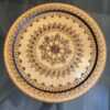 A round wooden tray features intricate, symmetrical floral and geometric patterns etched in dark brown. The center has a red and black flower, surrounded by multiple decorative borders and motifs. The tray rests on a glass table with a blurred background.