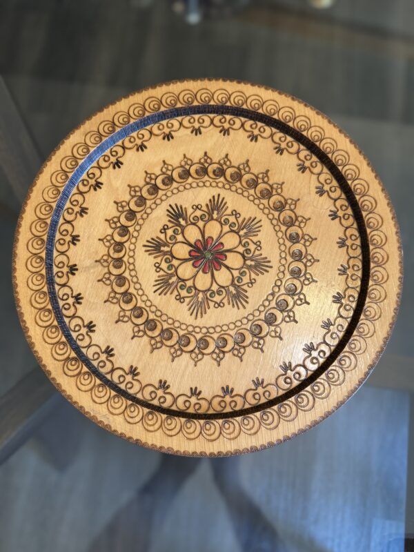 A round wooden tray features intricate, symmetrical floral and geometric patterns etched in dark brown. The center has a red and black flower, surrounded by multiple decorative borders and motifs. The tray rests on a glass table with a blurred background.