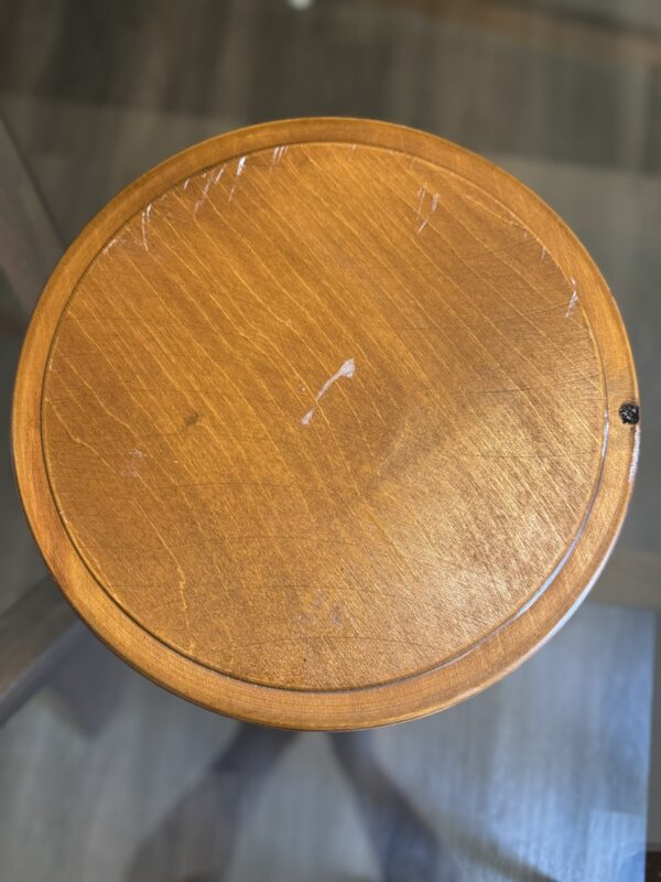 A round wooden stool with a warm brown finish is shown from above. The surface has visible scratches and marks, indicating wear. The background is out of focus, showing a dark wooden floor under the stool.
