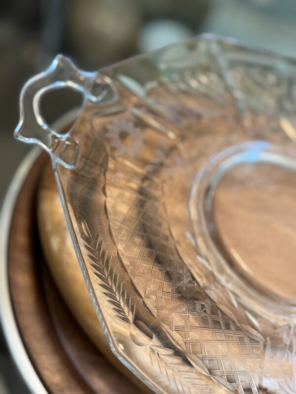 A close-up of a clear glass serving tray with an etched geometric and floral pattern, featuring a decorative handle. The tray rests on stacked wooden plates or cutting boards, with a softly blurred background.