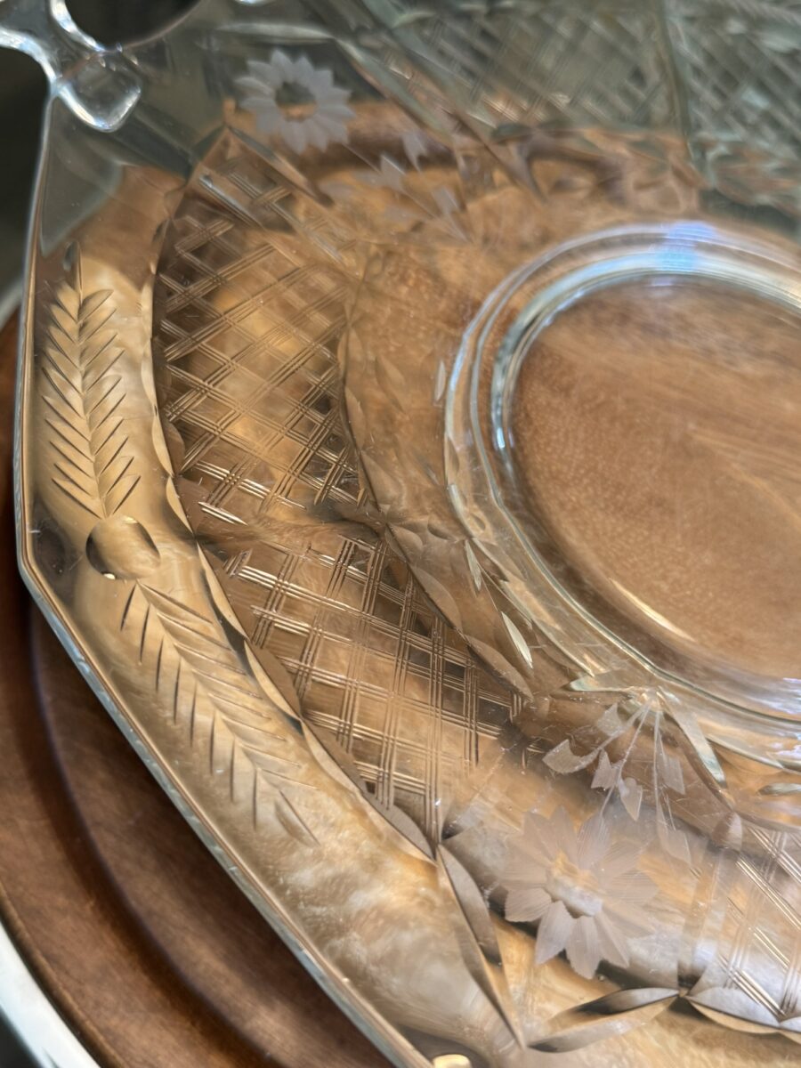 A close-up of a clear, intricately etched glass platter with floral and crisscross patterns along the rim, placed on a round wooden surface. The glass reflects light, highlighting its detailed design.