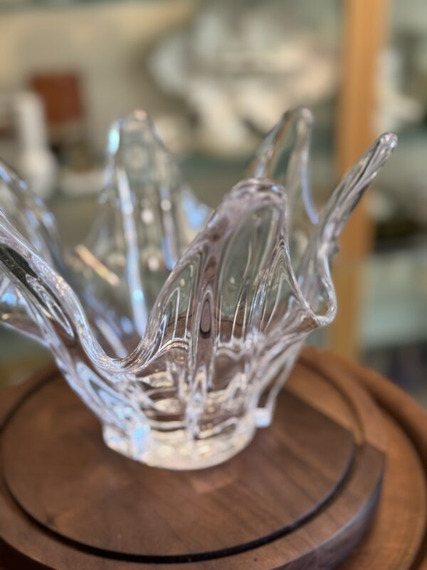 A clear glass decorative bowl with wavy, flared edges resembling flower petals sits on a round wooden surface. The background is softly blurred, showing shelves with various objects and contributing to a warm, inviting ambiance.