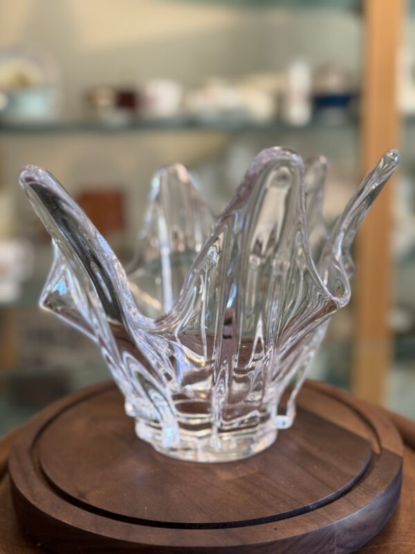 A clear glass decorative bowl with wavy, irregular edges resembling flames or petals sits on a round wooden rotating display. In the blurred background, shelves hold various small ceramic and glass items.