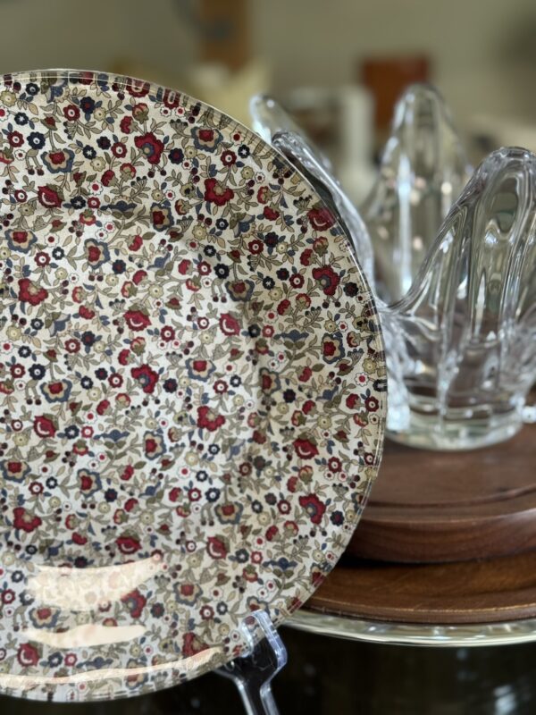 A round plate with a dense floral pattern in red, black, and beige sits on a clear stand. Behind the plate is a clear glass citrus juicer on a wooden surface, with blurred background objects.