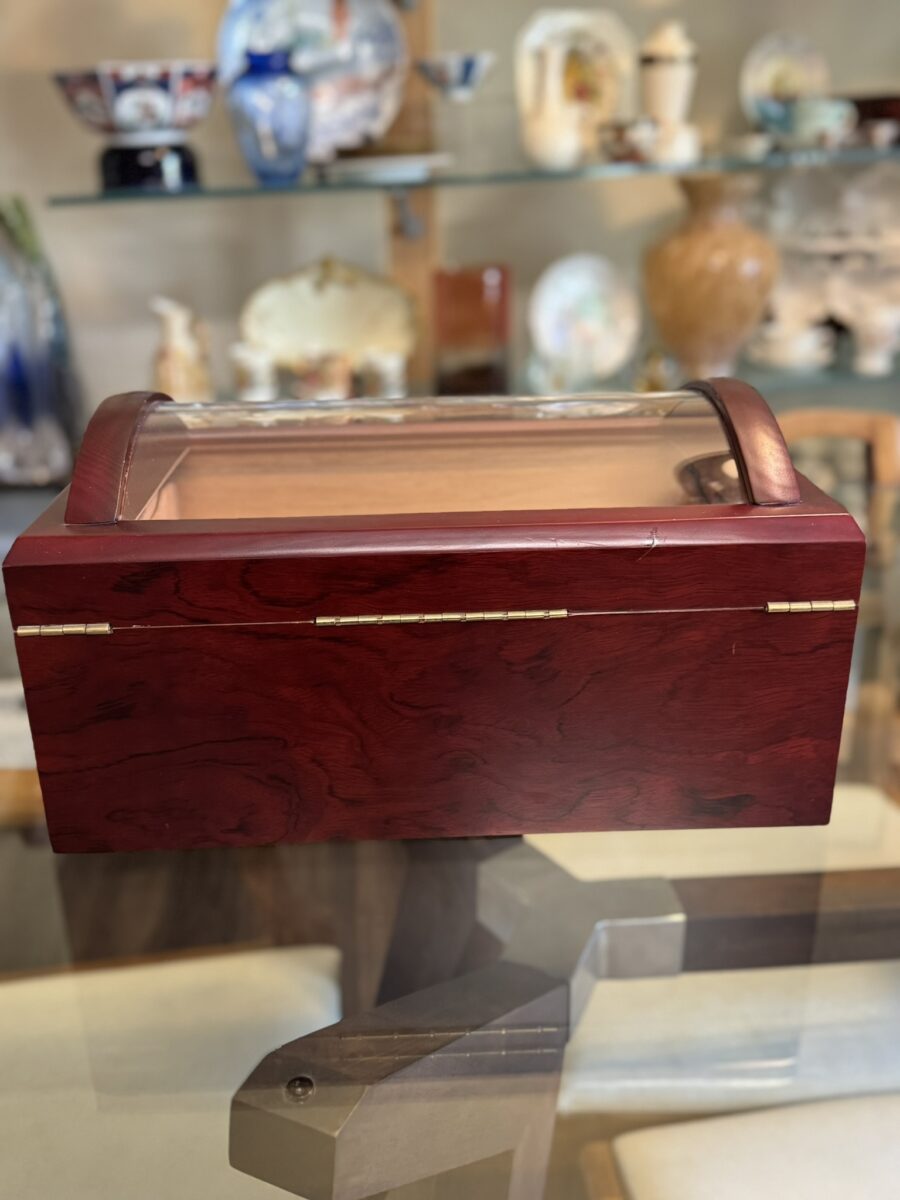 A polished wooden humidor with a glass top lid sits on a glass table. Behind it, shelves display various decorative porcelain vases, jars, and figurines with different colors and patterns, creating an elegant and eclectic background.