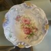 Ornate ceramic bowl with embossed swirls and gold accents on the rim, featuring a floral design with pink roses, white flowers, and green leaves in the center. The bowl is on a glass table with beige upholstered chairs visible below.