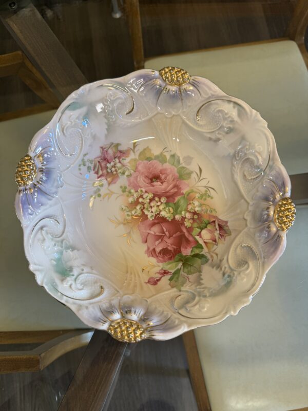 Ornate ceramic bowl with embossed swirls and gold accents on the rim, featuring a floral design with pink roses, white flowers, and green leaves in the center. The bowl is on a glass table with beige upholstered chairs visible below.