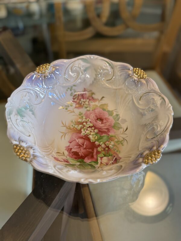 A decorative porcelain bowl with ornate gold accents on the rim and a detailed floral design of pink roses and white flowers in the center. The bowl sits on a glass table, with wooden furniture visible in the background.