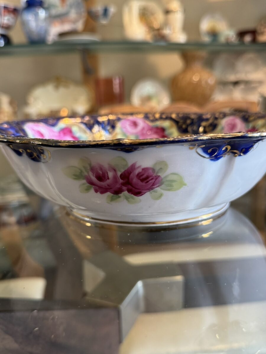 A decorative porcelain bowl featuring hand-painted pink roses and green leaves, with a dark blue scalloped rim adorned with gold accents. The bowl is displayed on a glass surface, with other ceramic items blurred in the background.
