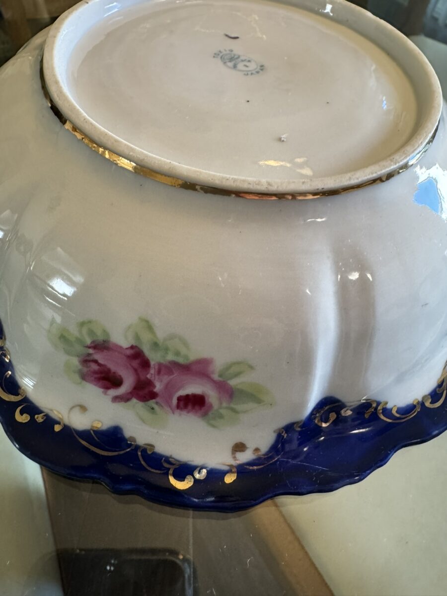 A close-up of the bottom of an ornate ceramic bowl, featuring hand-painted pink roses with green leaves on white porcelain, a blue border with gold decorative accents, and a faint maker’s mark on the base. The bowl rests upside-down on a glass surface.