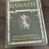A vintage book titled Hiawatha by Henry Wadsworth Longfellow is shown with a green cover, decorative borders, and a central illustration of crossed arrows. The text notes illustrations by Harrison Fisher. The book is resting on a dark wooden surface.