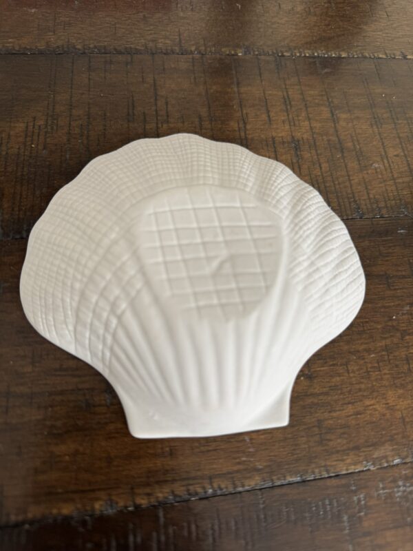 The Woulind Chile Serving Dish, a white ceramic scallop shell with textured ridges and a grid-patterned center, rests on dark wood, showcasing its detailed design and tactile appeal.