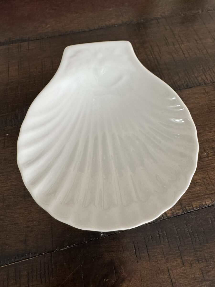 An Apilco Shell Dish, featuring a scallop shell design with smooth finish and detailed ridges, rests on a dark wooden surface, where the dish’s white porcelain contrasts with the rustic wood grain.