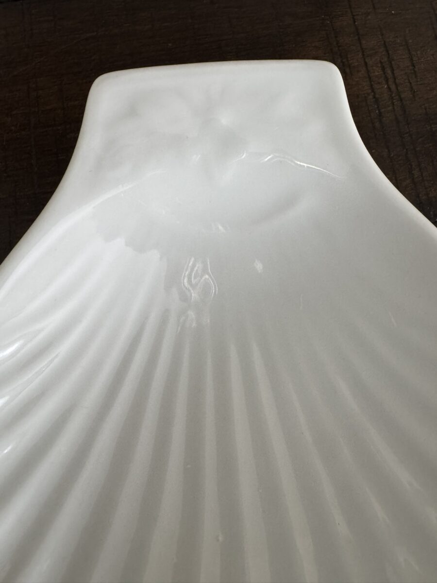 A close-up of the Apilco Shell Dish, shaped like a white scallop shell with gentle ridges and a subtle embossed bird near the top, displayed on a dark wooden surface.