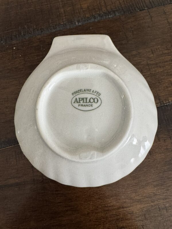An Apilco Shell Dish, made of white ceramic, rests upside down on a wooden surface, showing its scalloped edge and the maker’s mark 