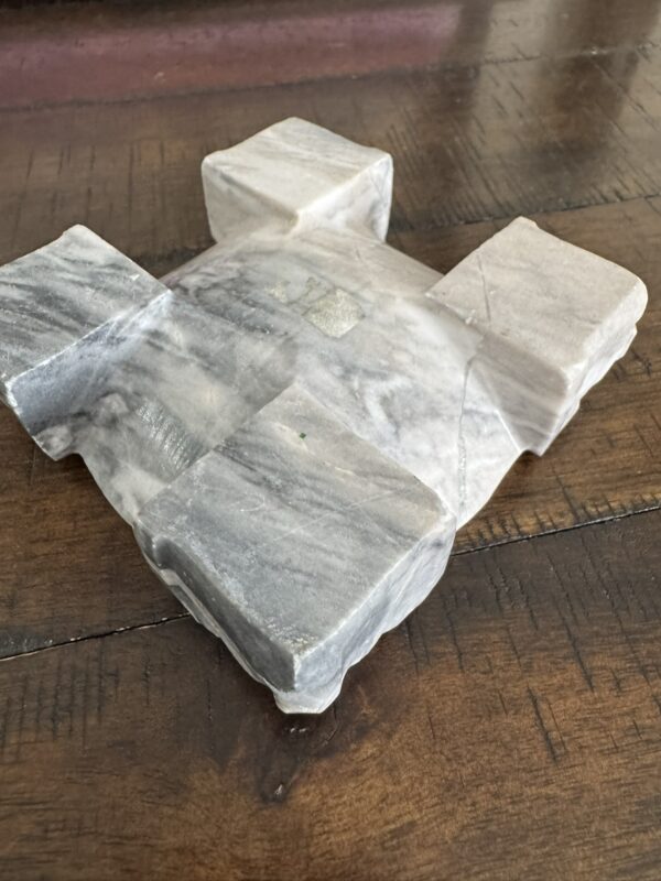 a marble cross-shaped piece with four square ends and a gray and white veined pattern, rests partially wrapped in translucent covering on a wooden surface, revealing some wood grain underneath.