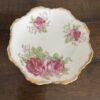 The Woulind Chile Serving Dish is a small porcelain piece with scalloped edges, gold trim, and a floral design of pink roses and green leaves. Three rose clusters are symmetrically arranged on its surface, which sits on dark wood.