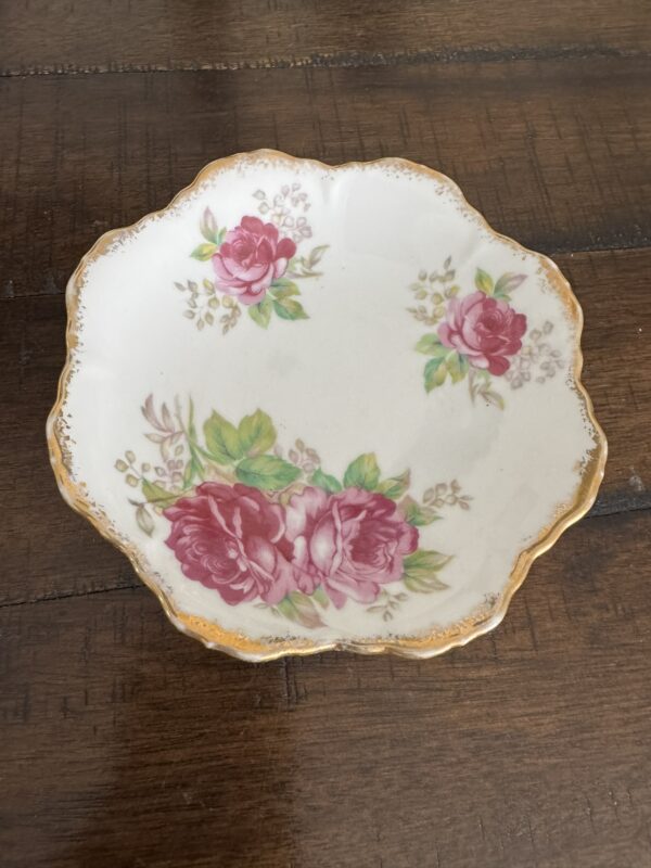 The Woulind Chile Serving Dish is a small porcelain piece with scalloped edges, gold trim, and a floral design of pink roses and green leaves. Three rose clusters are symmetrically arranged on its surface, which sits on dark wood.