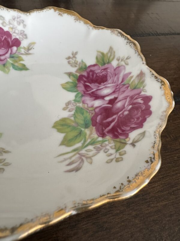 The Woulind Chile Serving Dish is an ornate ceramic piece with scalloped gold-trimmed edges and a floral design of pink roses, green leaves, and buds on a white background, displayed on a dark wooden surface.