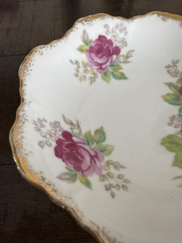 The Woulind Chile Serving Dish features painted pink roses, green leaves, and brown buds on white porcelain with scalloped gold-trimmed edges. Only part of the dish is visible in the image, set against a dark wooden surface.
