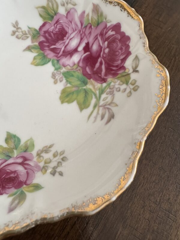 A close-up of the Woulind Chile Serving Dish, a porcelain piece with a scalloped gold-rimmed edge and floral design of pink roses and green leaves on white, displayed on a dark wooden surface.